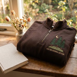 Peut inclure: Un sweat-shirt marron foncé à fermeture éclair quart, orné d'une broderie représentant trois pins verts, un croissant de lune et le texte "YOU'RE GONNA GO FAR" en lettres dorées sur la poitrine gauche, avec une fermeture éclair en métal.