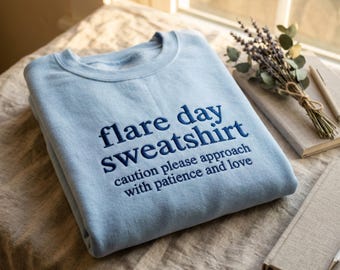 May include: A light blue crewneck sweatshirt featuring dark blue embroidered text that reads "flare day sweatshirt" followed by "caution please approach with patience and love" in a smaller font size, centered on the chest area.