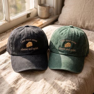 Puede incluir: Dos gorras de béisbol, una gris carbón y otra verde bosque, cada una con texto bordado que dice I just want some cheese and quiet junto a un pequeño gráfico de un trozo de queso amarillo centrado en los paneles frontales.