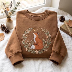Könnte beinhalten: Ein braunes Sweatshirt mit Rundhalsausschnitt und einer Stickerei, die einen Fuchs in einem kreisförmigen Kranz aus Blumen, Blättern und Stängeln zeigt. Über dem Fuchs sind eine kleine Mondsichel und mehrere kleine Sterne in gelbem Garn gestickt.