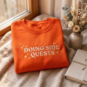 Könnte beinhalten: Ein leuchtend orangefarbenes Sweatshirt mit Rundhalsausschnitt und dem gestickten Text DOING SIDE QUESTS in cremefarbenen Großbuchstaben, akzentuiert durch kleine gelbe vierzackige Sternsymbole, die den Text auf der Vorderseite umgeben.