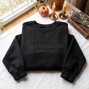 Könnte beinhalten: Ein schwarzes Langarm-Sweatshirt mit Rundhalsausschnitt und dem gestickten Text "THEY MISSED ONE" in Großbuchstaben, wobei die Worte "Salem, 1692" in einer kleineren, kursiven Schrift direkt unter dem Haupttext auf der Brust gestickt sind.
