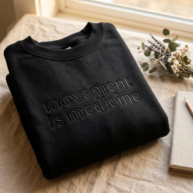 Op de afbeelding: Zwart sweatshirt met ronde hals met de geborduurde tekst "movement is medicine". Het sweatshirt is netjes op een neutraal gekleurd oppervlak gevouwen. Een notitieboek, potlood en takje gedroogde bloemen staan op de achtergrond.