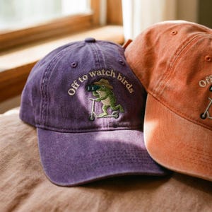 Könnte beinhalten: Eine violette Baseballkappe mit einer Stickerei eines grünen Frosches, der einen Hut trägt, ein Fernglas benutzt und auf einem Roller fährt, mit dem Text "Off to watch birds" darüber. Daneben ist ein Teil einer orangefarbenen Kappe zu sehen.