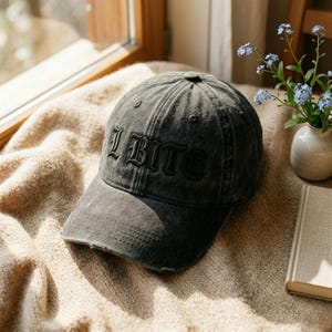 May include: A distressed black baseball cap with the word "I BITE" embroidered on the front. The cap has a curved brim and a worn appearance. The hat is sitting on a beige surface.