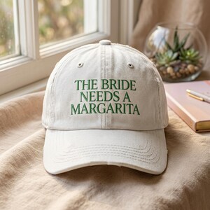 Może przedstawiać: Kremowa czapka bejsbolówka z zielonym haftem "THE BRIDE NEEDS A MARGARITA". Czapka ma zakrzywiony daszek i postarzany wygląd. Idealna na wieczór panieński lub imprezę ślubną.