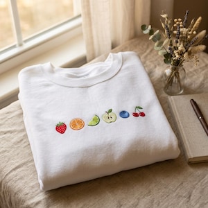 May include: White crew neck sweatshirt featuring embroidered fruit designs: strawberry, orange slice, lime, apple, blueberry, and cherries. The sweatshirt is folded on a neutral surface near a window.