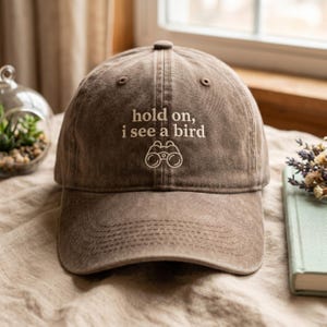 May include: Brown baseball cap with the embroidered text "hold on, i see a bird" and a pair of binoculars. The cap has a curved brim and a fabric strap closure. The hat is made of a soft, worn material.