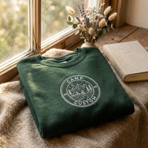 Op de afbeelding: Een donkergroene sweater met ronde hals en een witte cirkelvormige afbeelding op de borst. Het ontwerp toont een berglandschap met een tent, kampvuur, bomen en een maansikkel, omringd door de tekst "CAMP CUSTOM" en de cijfers "20" en "26".