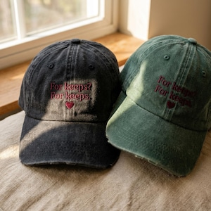 Peut inclure: Deux casquettes de baseball, une gris anthracite et une vert forêt, toutes deux dotées d'une finition en tissu délavé. Chaque casquette est brodée du texte "For keeps? For keeps." en fil rouge foncé, au-dessus d'une petite icône de cœur rouge.