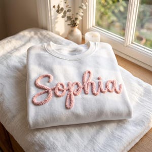 May include: A white crewneck sweatshirt featuring the name Sophia embroidered in a raised, textured, light pink script font across the chest. The garment has long sleeves and a ribbed neckline, showcasing a soft, plush finish on the lettering.