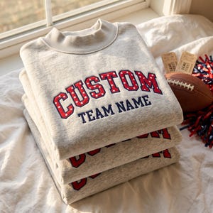 Op de afbeelding: Een stapel van drie lichtgrijze gemêleerde sweatshirts met ronde hals en geribbelde boorden. Het bovenste sweatshirt is voorzien van rood geborduurde tekst met witte stippen die CUSTOM spelt, met daaronder de woorden TEAM NAME in marineblauw.