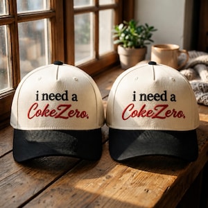 Puede incluir: Dos gorras de béisbol con coronas de color blanco roto y viseras negras. La parte delantera de cada gorra presenta el texto negro bordado "i need a" sobre el texto rojo bordado en cursiva "CokeZero." con un pequeño botón negro en la parte superior.