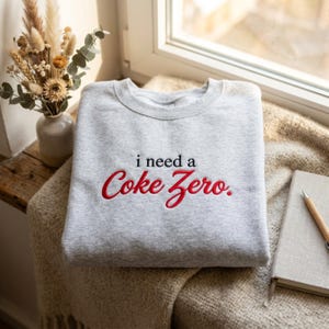May include: A light grey crewneck sweatshirt featuring the embroidered text i need a in black and Coke Zero. in red script. The garment has a classic long-sleeve design with a ribbed neckline, cuffs, and hem, showcasing a clean and simple aesthetic.