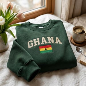 Può includere: Una felpa girocollo verde scuro con la parola GHANA ricamata in un carattere arcuato color crema sopra una piccola toppa rettangolare ricamata della bandiera nazionale del Ghana, che presenta strisce orizzontali rosse, gialle e verdi.
