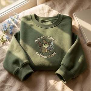 Może przedstawiać: Zielona bluza z napisem "OFF TO COMMIT SHENANIGANS" i grafiką żaby czarodzieja z kreskówek. Żaba ma na sobie kapelusz i trzyma laskę oraz książkę. Bluza jest złożona na beżowej powierzchni.