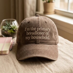 May include: Brown baseball cap with the embroidered text "I'm the primary breadloser of my household." The cap has a curved peak and a fabric strap closure. The hat is on a neutral-coloured surface.