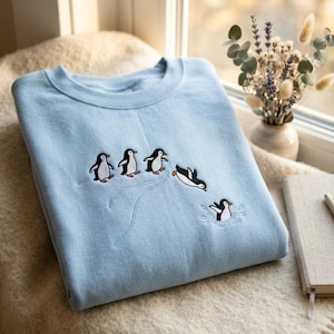 Op de afbeelding: Een lichtblauw sweatshirt met ronde hals en een geborduurd ontwerp van vijf pinguïns. Drie pinguïns staan op een lijn, één pinguïn maakt een duiksprong in de lucht en één pinguïn spat in het water, uitgevoerd in zwart, wit en oranje.