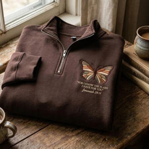 May include: A brown quarter-zip sweatshirt with a butterfly design and the text "FOR I KNOW THE PLANS I HAVE FOR YOU" Jeremiah 29:11. The butterfly is embroidered with shades of orange, brown, and green. The sweatshirt is on a wooden surface.