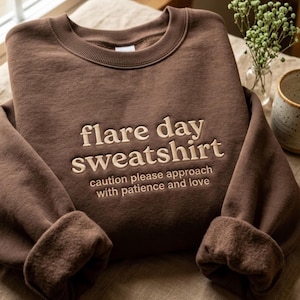 May include: A dark brown crewneck sweatshirt featuring beige embroidered text that reads: flare day sweatshirt, caution please approach with patience and love. The garment has long sleeves with ribbed cuffs and a ribbed neckline.