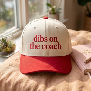 Könnte beinhalten: Cremefarbene Baseballkappe mit rotem Schirm und Knopf. Der Schriftzug "dibs on the coach" ist rot auf die Vorderseite gestickt. Ein lässiges Accessoire für Sportfans oder Trainer.