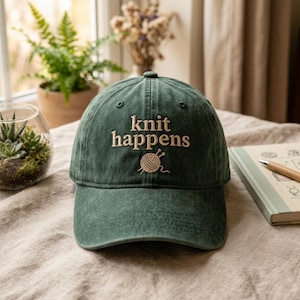 Op de afbeelding: Een groene baseballcap met de woorden "knit happens" geborduurd in een lichtbruine kleur. Een bol garen en naalden zijn onder de tekst geborduurd. De cap ligt op een tafel.