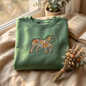 May include: A sage green crewneck sweatshirt featuring an embroidered design of a bulldog in profile. The dog's body is filled with a colorful pattern of orange flowers, green leaves, and purple sprigs, creating a detailed botanical illustration.