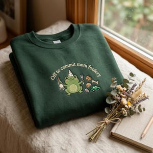 Könnte beinhalten: Ein dunkelgrüner Rundhals-Pullover mit einer Stickerei eines hellgrünen Frosches mit Partyhut. Der Frosch hält eine Flasche und eine Gabel, umgeben von kleinen Symbolen eines Kekses, Knochens, Autos und dem Text Off to commit mom foolery.