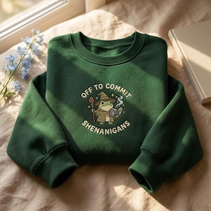 Puede incluir: Una sudadera de cuello redondo en color verde oscuro con un gráfico de una rana vestida con sombrero y túnica de mago, sosteniendo un bastón de madera y un libro abierto. El texto "OFF TO COMMIT SHENANIGANS" rodea la ilustración en arco.