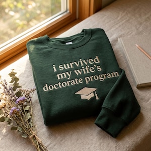 May include: A dark green sweatshirt with the embroidered text "i survived my wife's doctorate program" and a graduation cap. The sweatshirt is folded on a neutral-colored surface, with a book and pencil nearby.