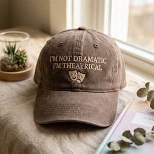 Peut inclure: Casquette de baseball marron avec la broderie "I'M NOT DRAMATIC I'M THEATRICAL" et deux masques de théâtre. La casquette est en matière souple et possède une visière incurvée. Une petite plante succulente dans un récipient en verre est visible en arrière-plan.