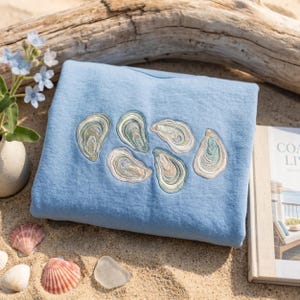 May include: A light blue sweatshirt featuring an embroidered pattern of six oyster shells in shades of green, tan, and white. The embroidery is centered on the front of the garment, showcasing detailed textures and natural shell color variations.
