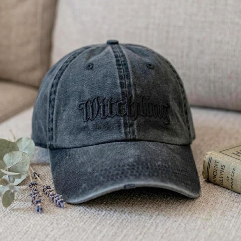 May include: A distressed charcoal gray baseball cap with the word "Witchling" embroidered in a matching dark thread. The cap has a curved brim and a classic six-panel design. The hat is sitting on a neutral-toned surface.