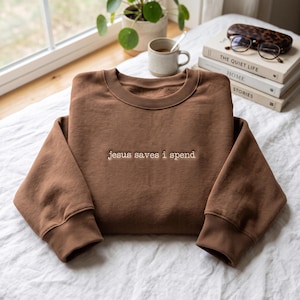 May include: A solid brown crewneck sweatshirt featuring the embroidered text "jesus saves i spend" in white thread across the centre chest. The garment has long sleeves with ribbed cuffs and a ribbed neckline, presented in a clean, folded layout.