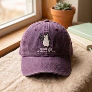 以下が含まれることがあります： ライフルを持ったペンギンの刺繍が施された紫色の野球帽。ペンギンの下には「FLIGHT WAS NEVER AN OPTION」という文字が刺繍されています。この帽子は構造化されたクラウン、カーブしたつば、そして色あせたような風合いの生地が特徴です。