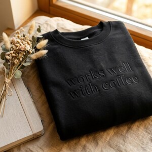 May include: Black crewneck sweatshirt with the phrase "works well with coffee" embroidered on the front. The sweatshirt is displayed on a neutral surface next to a notebook and a small bouquet of dried flowers.