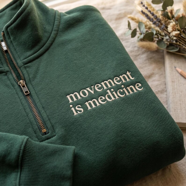 K&ouml;nnte beinhalten: Ein dunkelgr&uuml;nes Sweatshirt mit einem Viertel-Rei&szlig;verschluss-Ausschnitt und dem gestickten Text "movement is medicine" in Wei&szlig;. Das Sweatshirt hat eine gerippte Manschette und einen bronzefarbenen Rei&szlig;verschluss. Der Hintergrund umfasst ein Notizbuch, einen Bleistift und Trockenblumen.