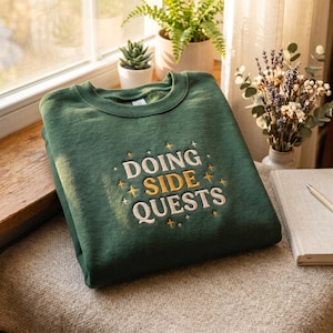 May include: A dark green crewneck sweatshirt featuring the embroidered text "DOING SIDE QUESTS" in white and gold thread, accented with small embroidered star shapes surrounding the lettering, displayed on a textured surface.