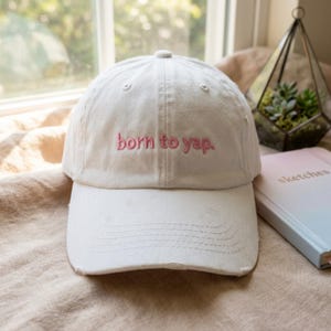 May include: A white baseball cap with a distressed brim and the text "born to yap." embroidered in pink thread across the front panel. The cap features a six-panel construction with a button on top and matching white stitching throughout the design.