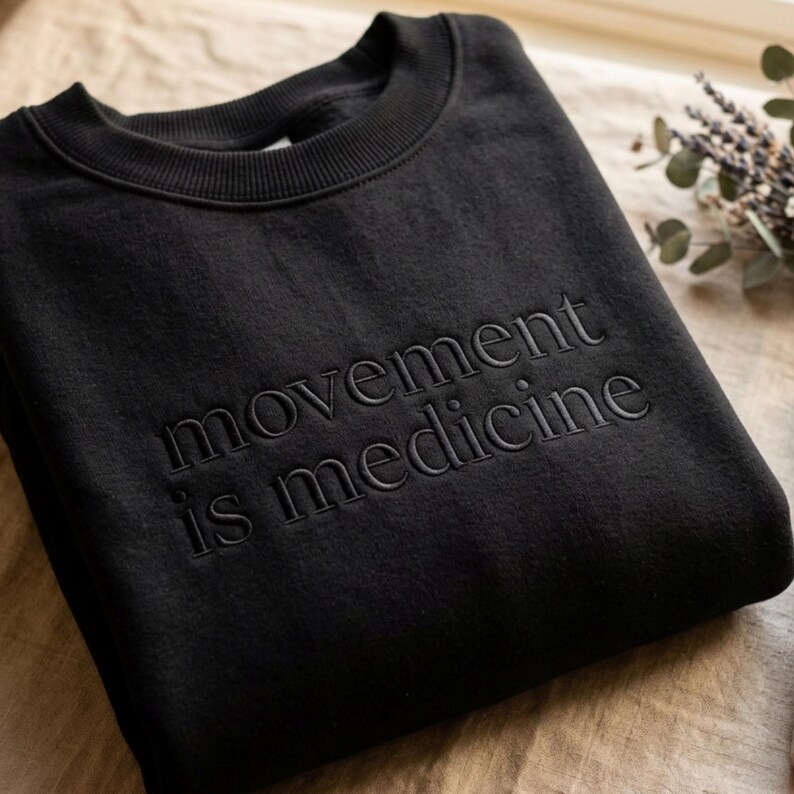 Op de afbeelding: Zwart sweatshirt met ronde hals en de geborduurde tekst "movement is medicine". Het sweatshirt is opgevouwen en lijkt van een zachte stof te zijn. De foto is genomen vanuit een iets verhoogde hoek.