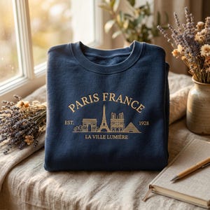 Peut inclure: Sweat-shirt bleu marine avec texte doré et un dessin de monuments parisiens, dont la Tour Eiffel et l'Arc de Triomphe. Le texte indique "Paris France" et "La Ville Lumière". Le sweat-shirt est plié sur une table.