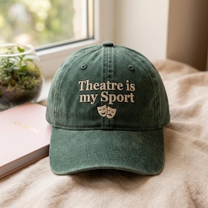Peut inclure: Casquette de baseball vert foncé avec l'inscription "Theatre is my Sport" et deux masques de théâtre brodés sur le devant. La casquette est en tissu doux et possède une visière incurvée. Un journal rose et une petite plante sont en arrière-plan.