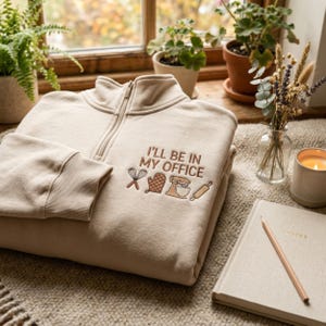 May include: A beige quarter-zip sweatshirt featuring brown embroidered text that reads I'LL BE IN MY OFFICE, accompanied by small stitched icons of a whisk, an oven mitt, a stand mixer, and a rolling pin, all centered on the chest area.
