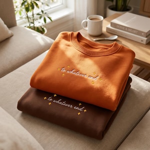 Pode incluir: Duas camisolas de gola redonda empilhadas uma sobre a outra. A camisola de cima é de cor laranja queimado com texto bordado a branco que diz "to whatever end" rodeado por pequenas estrelas amarelas. A de baixo é castanho escuro.