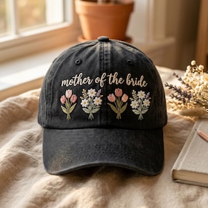 Puede incluir: Gorra de béisbol negra con la frase bordada "mother of the bride". Debajo, hay ramos de flores bordados en rosa, blanco y verde. La gorra está hecha de una tela suave y desgastada.