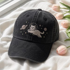 Könnte beinhalten: Schwarze Baseballkappe mit einem gestickten Design einer Katze in einem Weltraumhelm, Planeten und Sternen. Die Kappe ist aus einem gewaschenen, verblichenen Stoff.