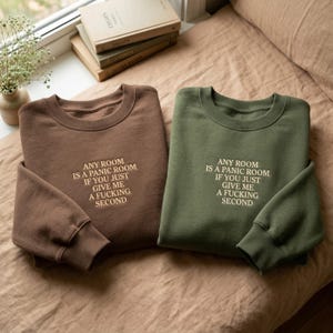 Könnte beinhalten: Zwei Sweatshirts mit Rundhalsausschnitt, eines in Braun und eines in Olivgrün, werden gezeigt. Jeder hat den gestickten Text: "ANY ROOM IS A PANIC ROOM IF YOU JUST GIVE ME A FUCKING SECOND." Die Sweatshirts liegen auf einer neutralfarbenen Oberfläche.