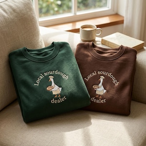 Könnte beinhalten: Zwei Rundhals-Sweatshirts, eines in Dunkelgrün und eines in Braun, jeweils mit einer Stickerei einer Gans, die eine Kochmütze und eine Schürze trägt und ein Brot hält, mit dem Text Local sourdough dealer um die Illustration herum.