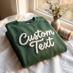 May include: A forest green crewneck sweatshirt featuring white, textured, raised chenille-style lettering that reads Custom Text across the chest. The garment has a ribbed collar, long sleeves, and a soft, fleece-lined interior fabric construction.