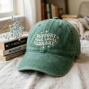 Peut inclure: Casquette de baseball verte avec le texte "SUPPORT YOUR LOCAL LIBRARY" brodé en blanc. La casquette a une visière incurvée et un design classique à six panneaux. Le chapeau est posé sur une surface de couleur neutre.